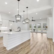 This image features a spacious kitchen with a large island and white shiplap detailing, topped with a marble-like countertop and a modern black faucet. Above the island, two unique square-framed pendant lights hang, adding character to the space.