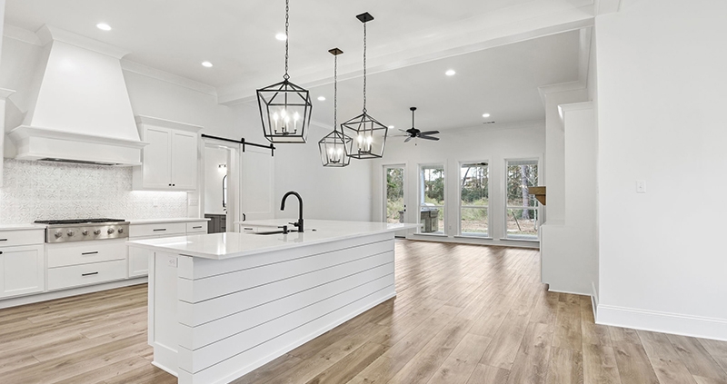 This image features a spacious kitchen with a large island and white shiplap detailing, topped with a marble-like countertop and a modern black faucet. Above the island, two unique square-framed pendant lights hang, adding character to the space.
