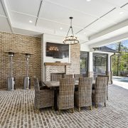 The image showcases an elegant outdoor dining area with a brick floor, featuring a large wooden dining table surrounded by wicker chairs, and a rustic chandelier overhead.