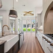 This kitchen captures the perfect blend of luxury craftsmanship and open-air beauty. A large central island with a stunning gray stone countertop features a deep farmhouse sink, sleek brushed gold faucet, and built-in dishwasher—ideal for prepping meals while enjoying the surrounding elegance. Overhead, modern black-and-gold pendant lights add a refined statement. To the right, a professional-grade gas cooktop is nestled beneath a dramatic arched plaster hood, creating an architectural focal point that’s both rustic and refined. The custom white cabinetry paired with polished hardware completes the gourmet aesthetic, while double ovens and ample counter space add functionality.