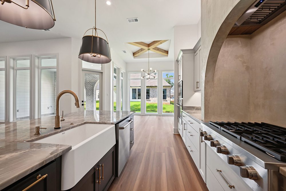 11-5040 Twin River Spacious Kitchen Design This kitchen captures the perfect blend of luxury craftsmanship and open-air beauty. A large central island with a stunning gray stone countertop features a deep farmhouse sink, sleek brushed gold faucet, and built-in dishwasher—ideal for prepping meals while enjoying the surrounding elegance. Overhead, modern black-and-gold pendant lights add a refined statement. To the right, a professional-grade gas cooktop is nestled beneath a dramatic arched plaster hood, creating an architectural focal point that’s both rustic and refined. The custom white cabinetry paired with polished hardware completes the gourmet aesthetic, while double ovens and ample counter space add functionality.