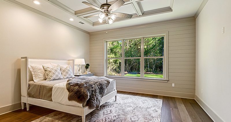The master bedroom has hardwood floors and a shiplap accent wall. The room is open and bright and features a coffered ceiling.