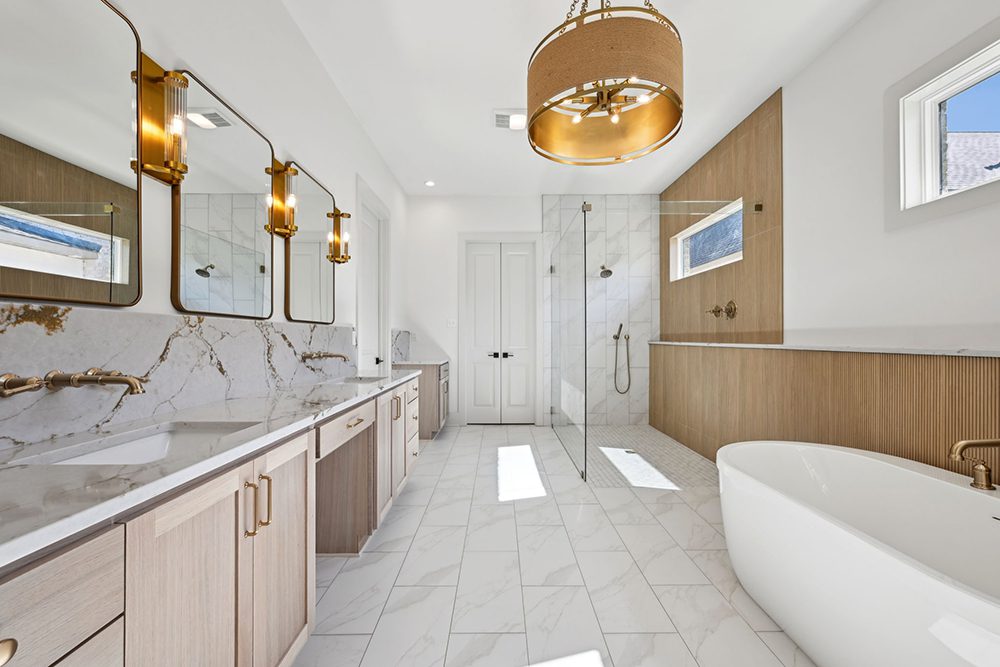 15-5040 Twin River Primary Bath Overview Overhead, a statement light fixture with a fabric and gold drum design enhances the spa-like ambiance. Bright natural light pours in through multiple windows, highlighting the clean lines and serene color palette of white, gold, and light wood. Every detail in this bathroom is thoughtfully curated to create a calming and luxurious retreat for everyday indulgence.