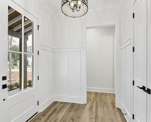 This image shows the interior of an entryway in a house, featuring white walls with wainscoting, a light fixture on the ceiling, and a view of the front door with glass panels.