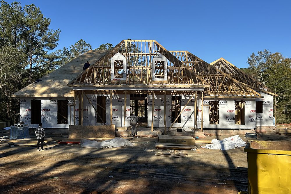 Folsom Custom Home Under Construction This photo captures a home under construction, showcasing its framework and roof structure in progress. The exterior walls are wrapped in Tyvek house wrap for weatherproofing, while exposed wooden trusses outline the roof and dormer windows.