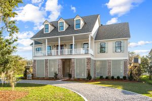 The lower level features latticework, adding both privacy and aesthetic appeal. A gravel driveway curves gracefully toward the entrance, creating a welcoming approach to the home. The lush greenery and well-maintained landscaping enhance the overall curb appeal, giving the home a sophisticated yet inviting feel.